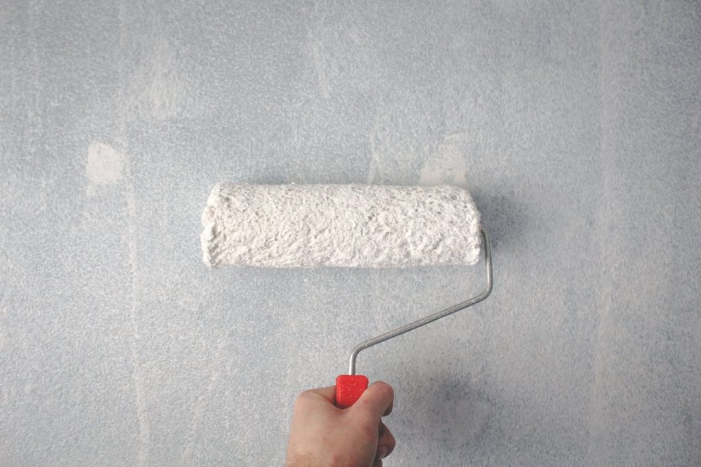A persons hand using a paint roller to apply white paint on a wall.